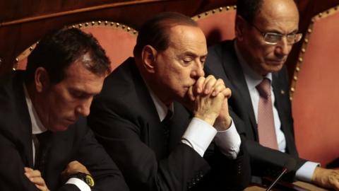 Mr Berlusconi sat with his hands crossed, flanked by two men