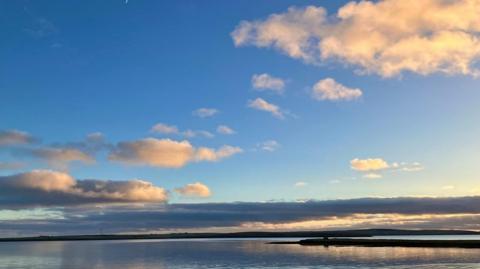 Some fluffy orange-tinged clouds float in a bright blue sky over the sea