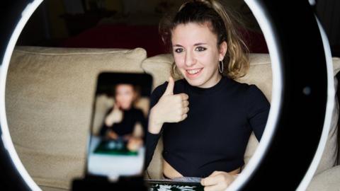 A young woman, framed by a circular ring light, sits on a sofa giving a thumbs up. In front of her, and in the middle of the ring light, a phone in vertical orientation records the scene. She's got her hair tied back and is smiling. She wears a black sweater and has large gold hoop earrings.