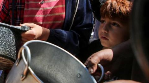 A young boy stands among a crowd holding bowls at an aid kitchen
