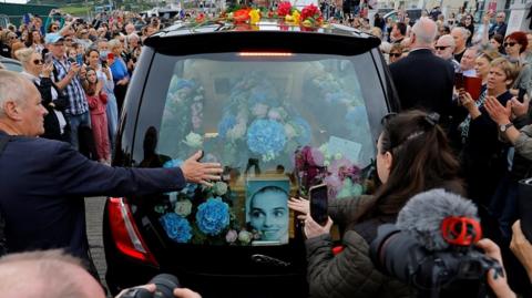 Hearse with crowds around it. A picture of Sinead O'Connor in the back