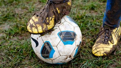photograph shows muddy football boots with one foot resting on a football