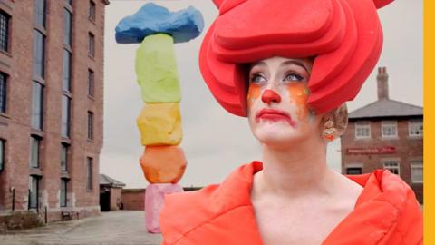 A woman dressed as a clown stands in front of Tate Liverpool staring into the distance