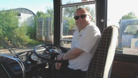 Steve Butler in the driver's seat of a bus smiling, wearing sunglasses