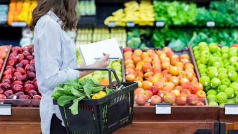A woman holds a notepad, pen and a shopping basket of vegetables. They are in a supermarket.