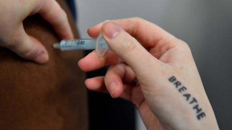 A Covid-19 booster vaccine injected into an arm by someone with the word breathe tattooed on their hand