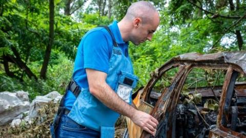 Kieran standing next to the shell of a burnt out car whilst wearing a safety vest.