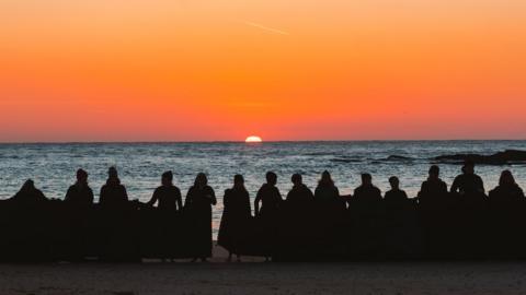 A group of women stand in front of the sea as the sun rises.