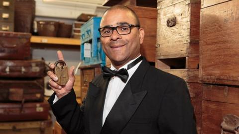 Shopowner, Carl, wearing a Bond style tuxedo and holding a golden padlock