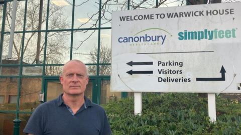 Man with short, light hair wearing blue top stands by Canonbury Products sign