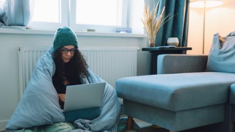 Women sits by radiator working from home wrapped in a duvet