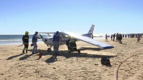 The plane body sits on the sand, under inspection from officials and surrounded by onlookers