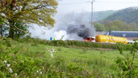 A photo of a fuel tanker on fire - thick smoke billows into the air. The surrounding area is very rural looking