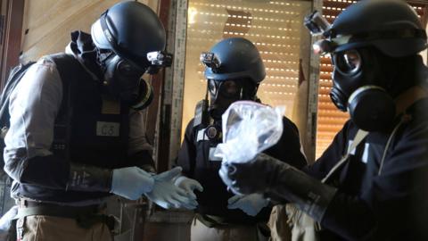 A UN chemical weapons expert, wearing a gas mask, holds a bag containing samples from a site of an alleged chemical weapons attack