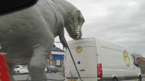 The dinosaur on the back of the trailer on the road