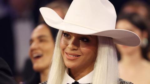 Beyoncé at the Grammys in January 2024. Beyoncé is a 42-year-old black woman with straight platinum blonde hair. She wears a white cowboy hat and white shirt with a studded black leather jacket.
