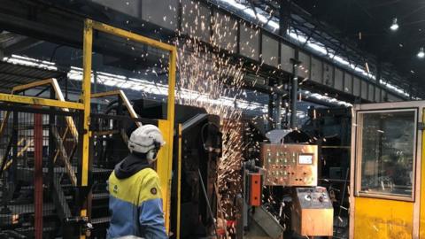 Sparks flying from machinery in a factory. A man in a hard hat is watching