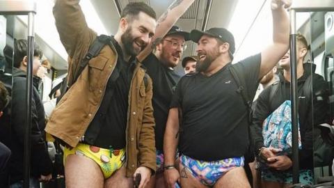Three men holding a handrail on a Tube train while wearing coconut and neon patterned pants.