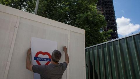 man puts up heart shaped grenfell poster in the shadow of the burnt out tower