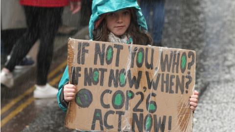 A child in a green coat holds a sign saying "If not you, who? If not now, when? COP26 act now!" at a protest in rainy Glasgow