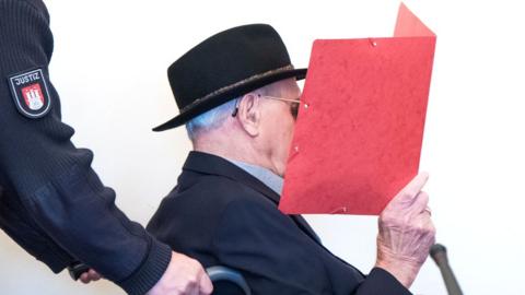 Mr Dey is seated in a wheelchair, wearing a black hat and sunglasses, as he holds a red folder up in front of his face