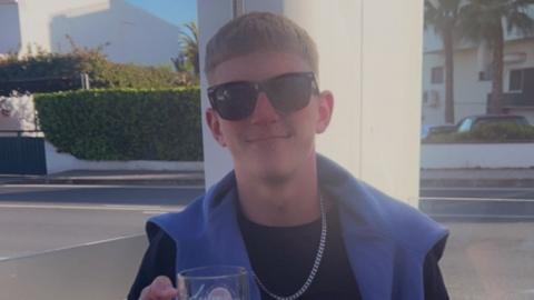 A young man with sunglasses on smiling at the camera wearing a black top and metal chain necklace