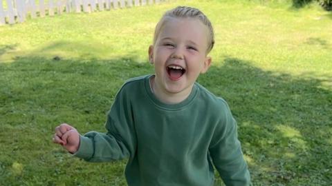 Happy girl wearing green top and blue trousers runs through a garden.