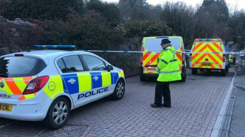 A female police officer wearing a high visibility jacket and standing by three police cars and a police cordon