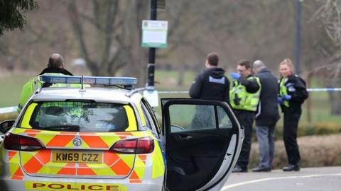 A police car in the foreground with officers and police tape in the background next to a grassy area of a park