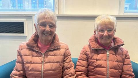 Two women with short white hair and glasses wearing identical pink coats