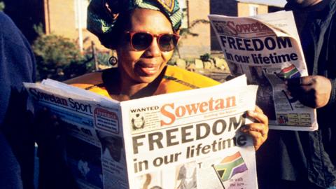 People line up to vote in the South Africa township of Soweto reading a paper with the headline "Freedom in our lifetime" - 27 April 1994