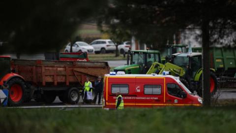 Rescuers stand by a firefighter vehicle, parked near the location where a farmer was killed and her husband and teenage daughter were seriously injured when they were run over at dawn on January 23, 2024