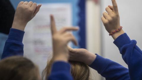 Young children with their hands up in a classroom. They they wearing dark blue jumpers and white shirts