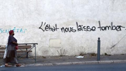 A woman walks in a street in "Les marronniers" neighbourhood as a handwritting reads 'State lets us down' on the wall, on August 30, 2021