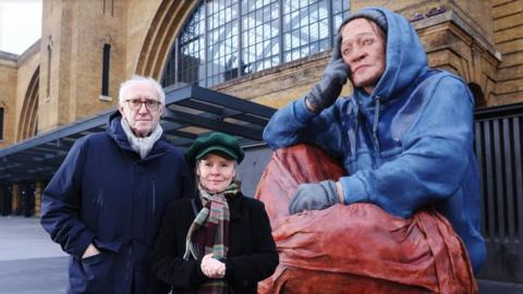 Imelda Staunton and Jonathan Pryce stand next to the sculpture, called Alex, outside King's Cross station.