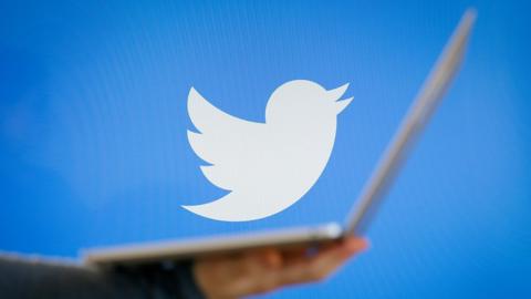 A Twitter logo (the white bird on a blue background) is seen in this photograph while a soft-focus foreground element shows a person's arm holding an open laptop