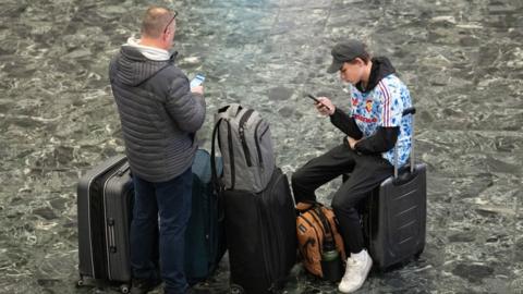 Two males sit on suitcases looking at their phone at a train station