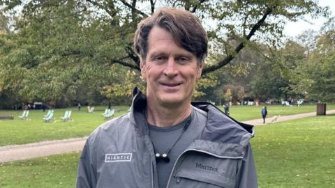 A middle-aged man with brown hair, parted at the side, stands in a park and smiles. He's wearing a grey t-shit underneath a thin, grey waterproof jacket which has a "Niantic" logo on the right breast and a "Marmot" logo on the left. He's got a pair of earbuds joined by a wire hanging from his neck. Behind him, we can see deck chairs, trees with green leaves just starting to show the first signs of autumn, and a woman walking her dog on a path that cuts through the park's grassed area.