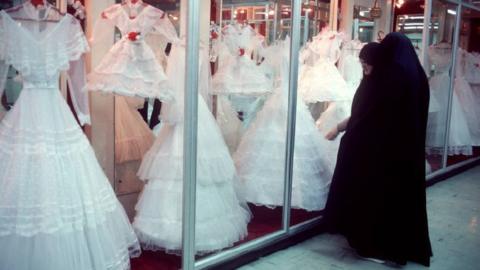 Two women wearing black chadors look at wedding dresses through glass windows in 1986