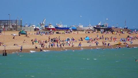 Green sea in front of a sandy beach full of people with boats lined up behind and a blue sky above