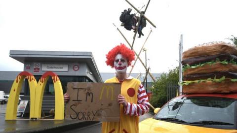 An Animal Rebellion protester, dressed as a clown, and protesters suspended from a bamboo structure outside a McDonalds distribution site in Hemel Hempstead