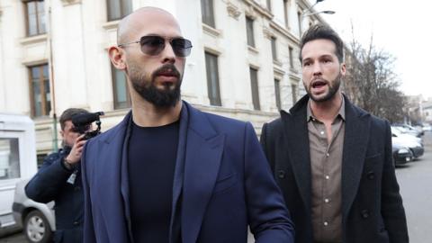 The Tate brothers appearing at court. Andrew is in the front with a bald head and sunglasses, and his brother Tristan is standing behind him