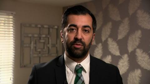 First Minister Humza Yousaf, wearing a suit with green tartan tie, stands in a room with silver leaf wallpaper.