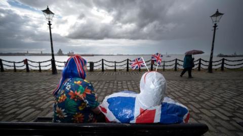 Two people clad with union jacks sit on a bench overlooking the river Mersey in Liverpool. Grey, rainy skies above and a person is walking by with an umbrella.