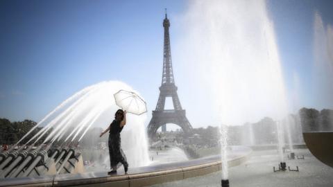 A woman in a summer dress holds a parasol over her head as the fountains in front of the Eiffel Tower spray her with a fine mist