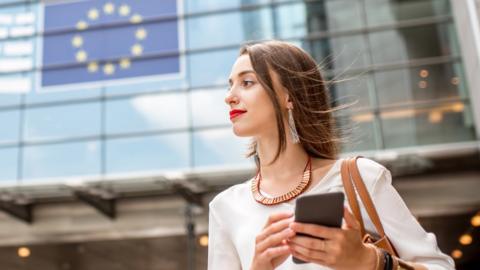 A stock image of a woman holding a phone in front of the EU parliament building