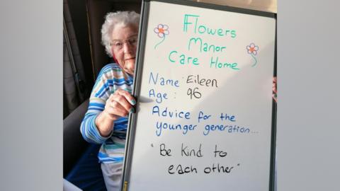 Eileen is wearing striped top and glasses, peeking out from behind the whiteboard with her message on