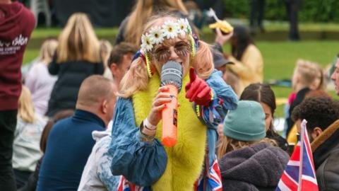 A woman holding an inflatable microphone and wearing fancy dress sings to the camera at Battersea Park.