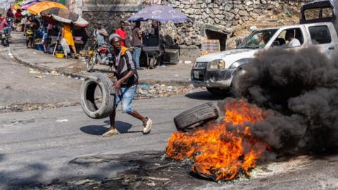Image shows unrest in Haiti as a man is about to throw a wheel into a pile of other burning car wheels.