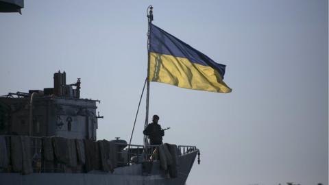 A Ukrainian naval ship, with a soldier standing in front of a Ukrainian flag